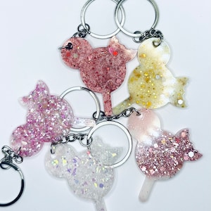 Peut inclure: Cinq porte-clés en résine pailletée de différentes couleurs en forme de sucettes. Chaque porte-clés a un motif de paillettes différent. Les porte-clés sont attachés à des anneaux de clés argentés.