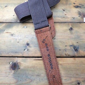 May include: A brown guitar strap with a herringbone pattern and cork backing. The strap has the text "DAD ROCKS" printed on it.