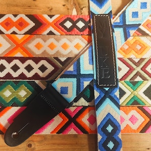 May include: A colourful guitar strap with a geometric pattern. The strap is made of woven fabric and has a black leather adjustment strap with the word "MYUKE" embossed on it.