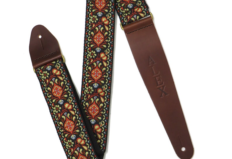 May include: A guitar strap with a black, red, and green floral pattern and a brown leather end with the word "ALEX" embossed on it.