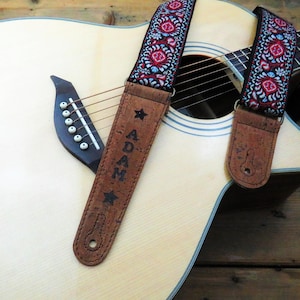 May include: A brown leather guitar strap with a blue and red patterned fabric. The strap has the name "ADAM" written on it in black lettering. The strap is attached to an acoustic guitar.