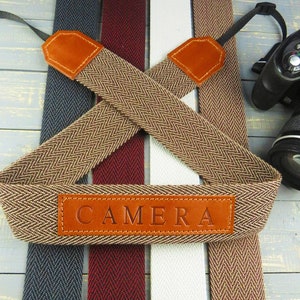May include: A brown camera strap with a herringbone pattern and leather accents. The strap has a leather patch with the word "CAMERA" embossed on it.
