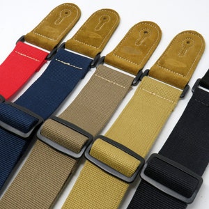 May include: Five guitar straps in different colours: red, navy blue, beige, khaki, and black. Each strap has a suede leather end with a keyhole design and a black metal buckle.
