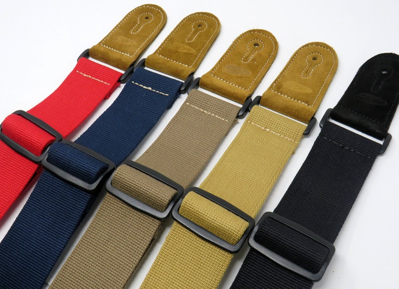 May include: Five guitar straps in different colours: red, navy blue, beige, khaki, and black. Each strap has a suede leather end with a keyhole design and a black metal buckle.