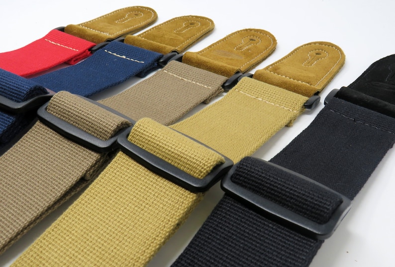 May include: Five guitar straps in different colours: red, blue, beige, tan, and black. Each strap has a black buckle and a brown suede end piece.
