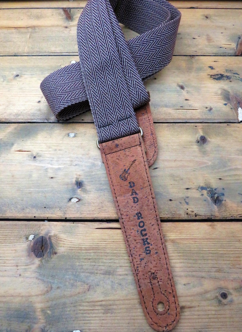 May include: A brown guitar strap with a herringbone pattern and cork backing. The strap has the text "DAD ROCKS" printed on it.