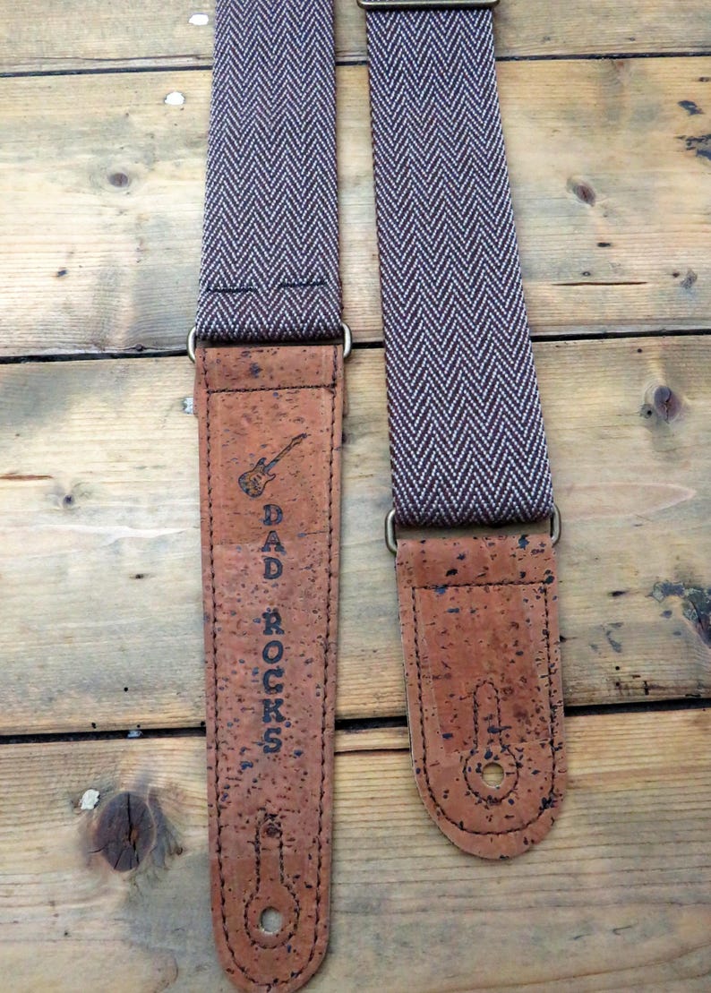 May include: A brown and white guitar strap with a cork end. The cork end has the text "DAD ROCKS" and a small guitar graphic.