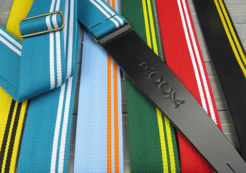 May include: A close-up of seven guitar straps with different colored stripes. The straps are made of fabric and have a leather end with the word "ROCK" embossed on it.