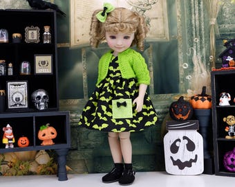 Puede incluir: Una muñeca con cabello rubio en coletas, vistiendo un cárdigan de punto verde lima sobre un vestido negro con estampado de murciélagos verde lima. El vestido incluye un bolsillo verde lima con un pequeño lazo negro. La muñeca lleva zapatos negros.