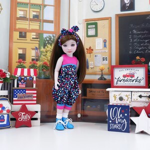 Op de afbeelding: Een pop in een outfit met sterrenpatroon, roze top en blauwe schoenen, staat voor patriottische decoraties. De scène omvat een bord met de tekst "All American Fireworks" en een "July 4th" ster, wat een feestelijke sfeer oproept.