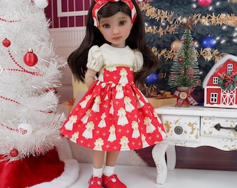 Puede incluir: Una muñeca con un vestido rojo con figuras de ángeles blancos, una diadema a juego y zapatos rojos. La muñeca tiene el pelo largo y castaño y está de pie frente a un árbol de Navidad y un árbol de Navidad blanco.
