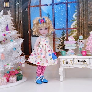 May include: A doll wearing a cream-colored dress with a colorful cupcake pattern, pink and white striped leggings, and blue shoes. The doll has blonde curly hair with a blue bow. A white Christmas tree and presents are in the background.