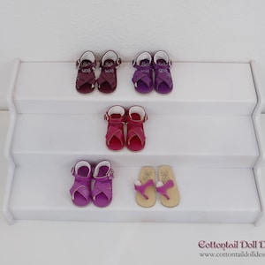 Puede incluir: Una estantería blanca escalonada exhibe varios pares de sandalias para muñecas. Las sandalias son de varios colores, incluyendo burdeos, morado y rosa. Algunas tienen cierres de hebilla, otras son tipo chanclas.