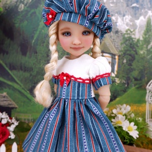 Op de afbeelding: Een pop die een blauw-wit gestreepte jurk draagt met witte sterren, een wit topje en een blauw-wit gestreepte hoed met een rode strik. De pop heeft blond haar in vlechten en blauwe ogen.
