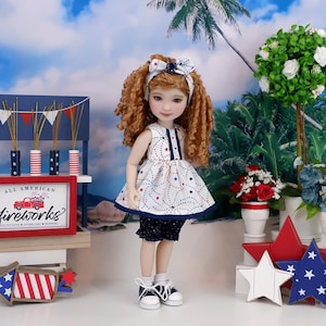Op de afbeelding: Een pop gekleed in een witte jurk met rode en blauwe sterrenpatronen, een bijpassende haarband en marineblauwe shorts. De pop staat voor een display met een bord met de tekst "All American Fireworks".