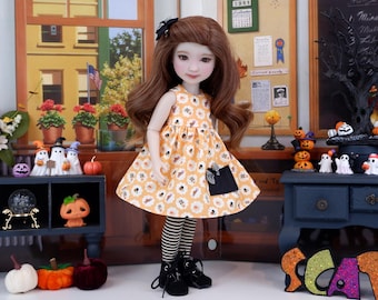 Puede incluir: Muñeca con un vestido naranja con estampado floral blanco y un lazo negro. La muñeca tiene cabello castaño, botas negras y leggings a rayas. Decoraciones de Halloween en el fondo.