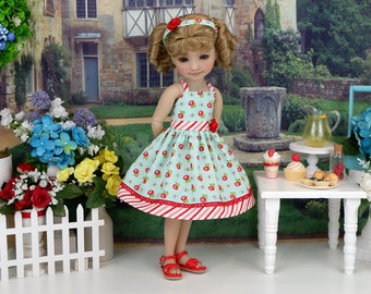 Puede incluir: Una muñeca con un vestido azul claro con una raya roja y blanca y un estampado de fresas. La muñeca tiene el pelo rubio y lleva sandalias rojas. La muñeca está de pie frente a una mesa blanca con una jarra de limonada y algunos postres.