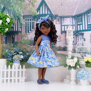 May include: A doll wearing a blue floral print dress with yellow accents, a matching headband, and blue sandals. The doll has dark hair and is posed in front of a house with a white picket fence and flowers.