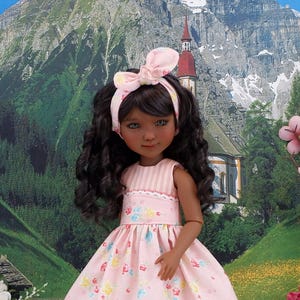 May include: A doll with dark brown curly hair and a light pink headband featuring a bow. The doll wears a sleeveless dress with a light pink striped bodice and a skirt patterned with small pink, blue, and yellow flowers on a light pink background.