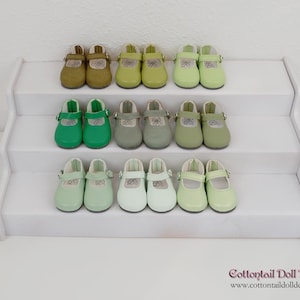 Peut inclure: Présentation de chaussures de poupée dans diverses nuances de vert, beige et blanc, disposées sur une étagère blanche à plusieurs niveaux. Les chaussures sont de style Mary Jane avec des fermetures à boucle. La gamme comprend plusieurs couleurs, du vert clair à l'olive.