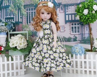 Puede incluir: Una muñeca con un vestido de estampado de margaritas blanco y negro, una diadema blanca y una flor blanca. La muñeca tiene el pelo largo, rojo y rizado y ojos azules. La muñeca está de pie frente a una valla blanca de madera.