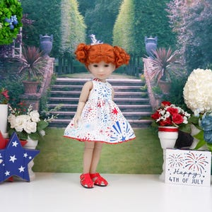 Op de afbeelding: Een pop met rood haar in een witte jurk met een rood, wit en blauw vuurwerkpatroon. De pop draagt rode sandalen. Een houten bordje zegt "Happy 4th of July".