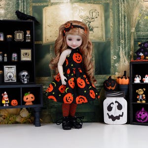 May include: A doll with long wavy brown hair wearing a black dress featuring an orange pumpkin and green star pattern, a matching headband, orange and black striped leggings, and black boots, standing in front of a Halloween-themed backdrop.