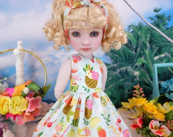 Puede incluir: Una muñeca con cabello rubio rizado y ojos azules que lleva un vestido azul claro con un estampado de frutas tropicales. El vestido presenta piñas, fresas y otras frutas. Una diadema a juego completa el look. El fondo incluye flores y un tucán.