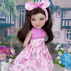 Puede incluir: Una muñeca con cabello largo castaño y ojos azules que viste un vestido rosa con estampado de hadas y una diadema rosa a juego con un lazo. El vestido presenta un corpiño a rayas y una falda fruncida con ilustraciones de hadas y flores.