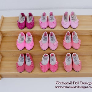 Puede incluir: Exhibición de bailarinas de muñeca en miniatura en varios tonos de rosa y fucsia, dispuestas sobre una estantería de madera escalonada. Cada par presenta un detalle de lazo y un diseño impreso en el interior. Incluye el texto "Cottontail Doll Designs".