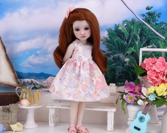 Puede incluir: Una muñeca con largo cabello castaño rojizo que lleva un vestido estampado y sandalias rosas. La muñeca posa en un banco blanco frente a un telón de fondo tropical. Un pequeño ukelele azul y flores están cerca.