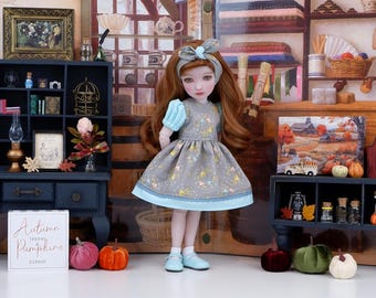 Puede incluir: Una muñeca con un vestido gris con ribete y mangas azul claro, y un lazo a juego. El vestido tiene un estampado de formas amarillas y azules. La muñeca está de pie frente a una escena en miniatura con calabazas y un letrero que dice "Autumn leaves & pumpkins please."