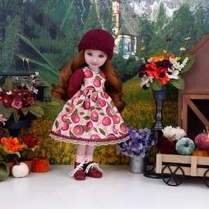 Puede incluir: Una muñeca con gorro de punto burdeos, zapatos a juego y un vestido con estampado de frutas rosas y rojas. La muñeca tiene el pelo largo pelirrojo y posa frente a un telón de fondo de un paisaje montañoso y flores.