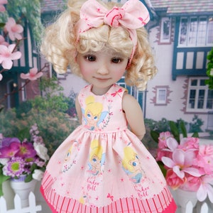May include: A porcelain doll with blonde curly hair wearing a pink dress with a Tinker Bell print and a pink bow headband. The doll is standing in front of a pink and green floral backdrop.