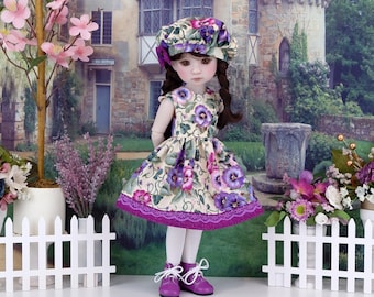 Puede incluir: Una muñeca con un vestido floral, sombrero a juego, medias blancas y zapatos morados. El vestido presenta flores moradas sobre un fondo crema, con un ribete de encaje morado. La muñeca tiene cabello castaño y está frente a un telón de fondo escénico.