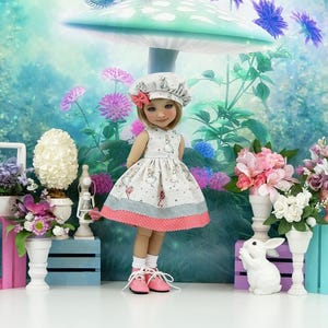 Puede incluir: Una muñeca viste un vestido beige sin mangas con un estampado de personajes repetido, un gorro a juego con un pequeño lazo rosa, calcetines blancos y zapatos rosas con cordones. El vestido presenta una sección gris y un dobladillo con lunares.