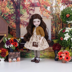 Può includere: Una bambola con un vestito floreale, cardigan color senape e leggings a righe. Indossa scarpe marroni e ha lunghi capelli castani. Un cartello con "Hello Fall" è visibile.
