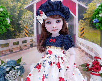 Puede incluir: Una muñeca de pelo castaño y ojos azules que lleva una boina azul oscuro a rayas finas con un lazo blanco, y un vestido con corpiño azul oscuro a rayas finas con un pequeño lazo rojo y falda color crema con estampado floral rojo y azul.