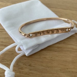 May include: Rose gold-coloured bracelet engraved with the name "SIENNA" and a heart symbol. The bracelet is presented on a white pouch with a drawstring. The background is a light-coloured wooden surface.