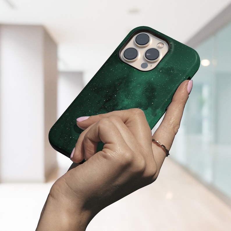 May include: A dark green phone case with a speckled, galaxy-like design. The case is held in a hand, showcasing the camera lenses and the case's texture. The case is designed for a smartphone.