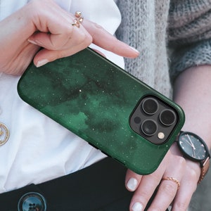 May include: A dark green phone case with a nebula design and a black camera module. The case has a glossy finish and a starry pattern. The phone case is held by a person.