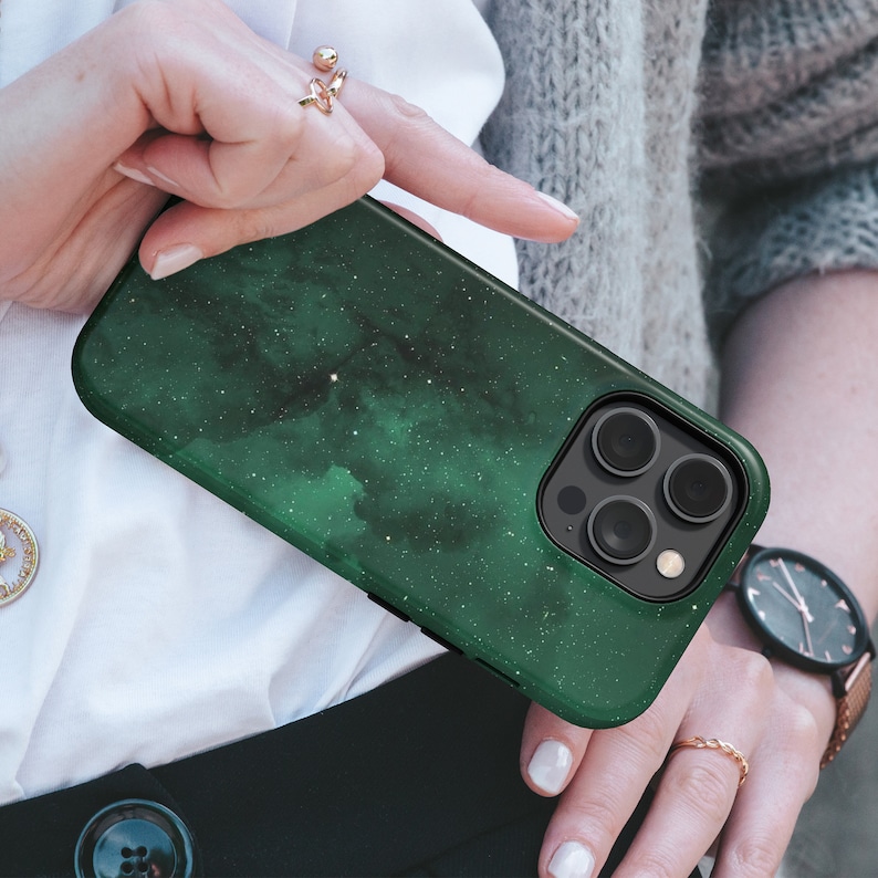 May include: A dark green phone case with a nebula design and a black camera module. The case has a glossy finish and a starry pattern. The phone case is held by a person.