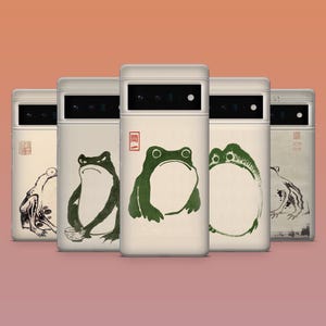 May include: A collection of five phone cases with a cream-colored base and a variety of Japanese-inspired frog illustrations in black and green. The cases feature a camera cutout and a minimalist design.