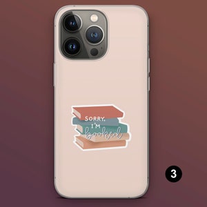 Puede incluir: Funda de tel&eacute;fono rosa claro con una pegatina de una pila de libros. La pegatina tiene el texto "Sorry, I'm booked" en una fuente cursiva. Los libros son en tonos marr&oacute;n, azul y verde.