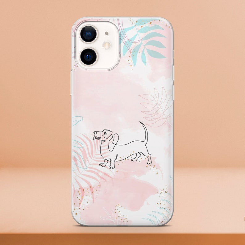 May include: Clear phone case featuring a line drawing of a dachshund dog. The case has a pink watercolor background with blue and gold leaf accents. The phone case is designed for an iPhone.