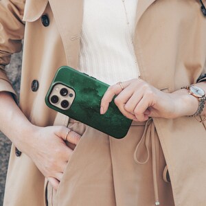 May include: A dark green phone case with a white speckled design, held in front of a tan trench coat. The phone case has a camera module with multiple lenses. The case has a textured, cloudy pattern.