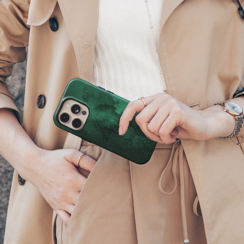 May include: A dark green phone case with a white speckled design, held in front of a tan trench coat. The phone case has a camera module with multiple lenses. The case has a textured, cloudy pattern.