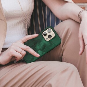 May include: A green phone case with a speckled design, held by a person. The case has a gold-colored camera ring and a dark green speckled pattern. The person is wearing rings and a gold necklace.