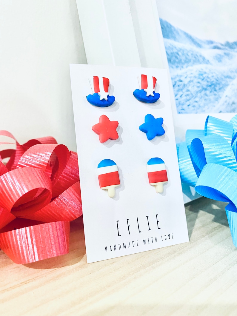 May include: A set of six red, white, and blue polymer clay earrings. The earrings are shaped like stars, ice cream cones, and top hats. The earrings are displayed on a white card with the text "EFLIE HANDMADE WITH LOVE".