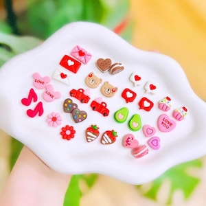 May include: A collection of colorful, miniature decorative items on a white, wavy-edged tray. The assortment includes hearts, strawberries, musical notes, bears, cars, avocados, cupcakes, and envelopes. Some items have the word "LOVE" or "LOVE YOU" on them.